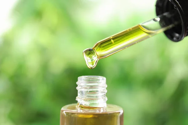 Dripping essential oil from pipette into glass bottle against blurred green background, closeup