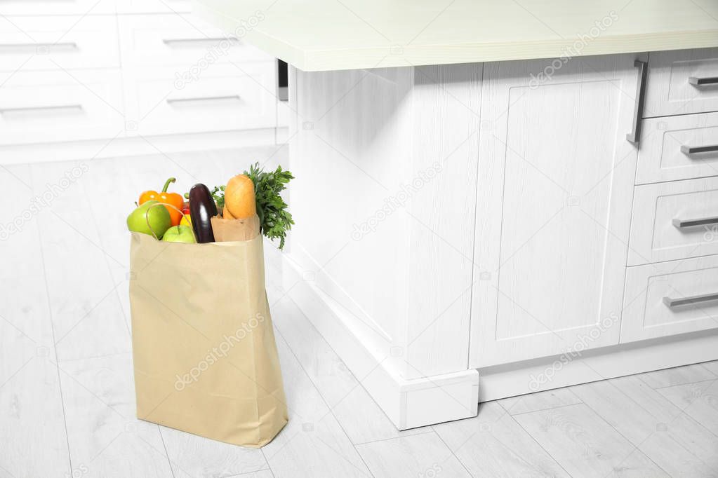 Paper shopping bag full of vegetables and baguette on floor in kitchen. Space for text