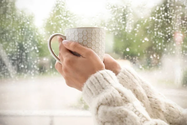Młoda kobieta z filiżanką herbaty w pobliżu okna wewnątrz w deszczowy dzień, zbliżenie — Zdjęcie stockowe