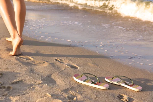 Primer plano de mujer y chanclas en arena cerca del mar, espacio para el texto. Accesorios de playa —  Fotos de Stock