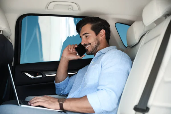 Atractivo joven que trabaja con el ordenador portátil y habla por teléfono en un coche de lujo — Foto de Stock