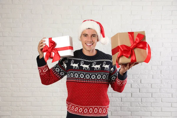 Happy man in Christmas sweater and Santa hat holding gift boxes near white brick wall — Stock Photo, Image