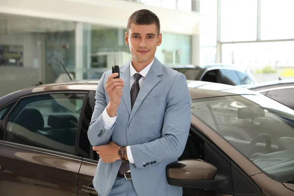 Jovem vendedor com chave perto do carro na concessionária moderna — Fotografia de Stock