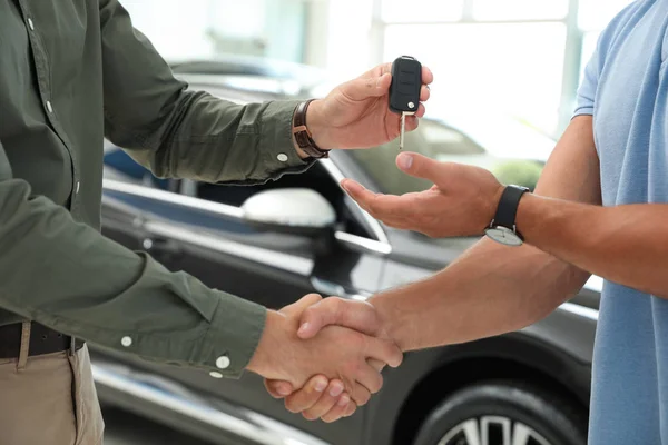 Młody sprzedawca potrząsając rękami z klientem i dając klucz samochodowy w nowoczesnym salonie samochodowym, zbliżenie — Zdjęcie stockowe