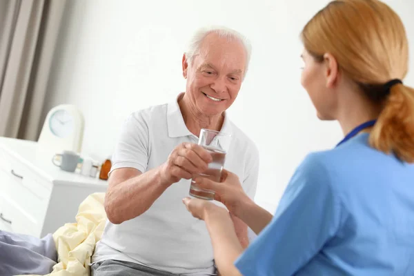 Enfermeira dando copo de água para o homem idoso dentro de casa. Assistência médica — Fotografia de Stock