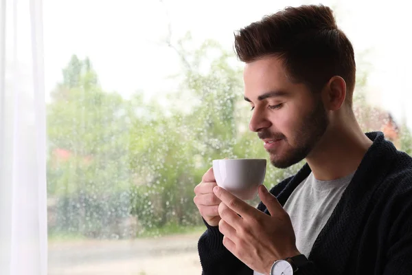 Boldog jóképű ember csésze kávé közelében ablak esős napon — Stock Fotó