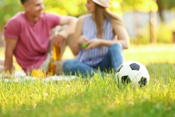 Vista borrosa de la pareja joven haciendo un picnic en el soleado parque de verano, se centran en la pelota de fútbol — Foto de Stock