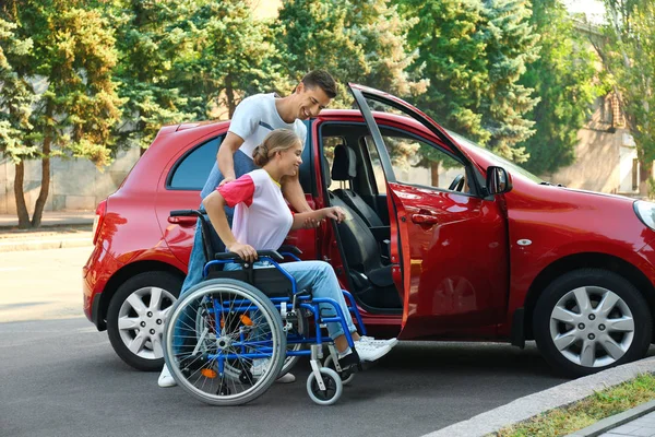Молодой человек помогает женщине-инвалиду в инвалидном кресле садиться в машину на открытом воздухе — стоковое фото