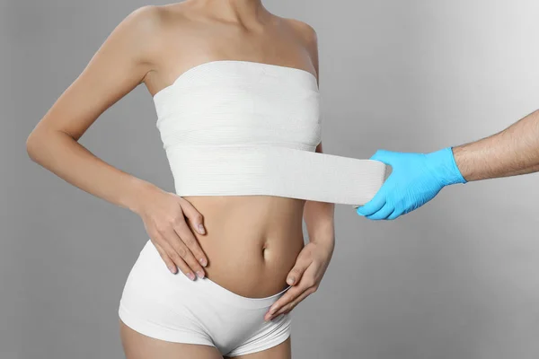 Doktor bandování mladé ženy po plastických operacích na šedém pozadí, zaostřená — Stock fotografie
