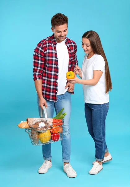 Fiatal pár, bevásárlókosárral, tele termékekkel kék háttérrel — Stock Fotó