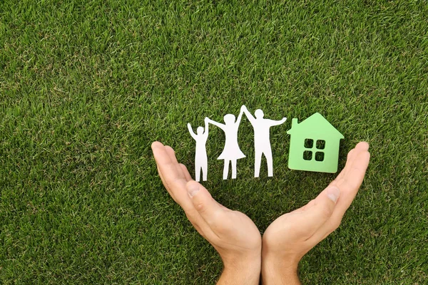 Мужчина, держащийся за руки возле фигур дома и семьи на зеленой траве, вид сверху. Пространство для текста — стоковое фото