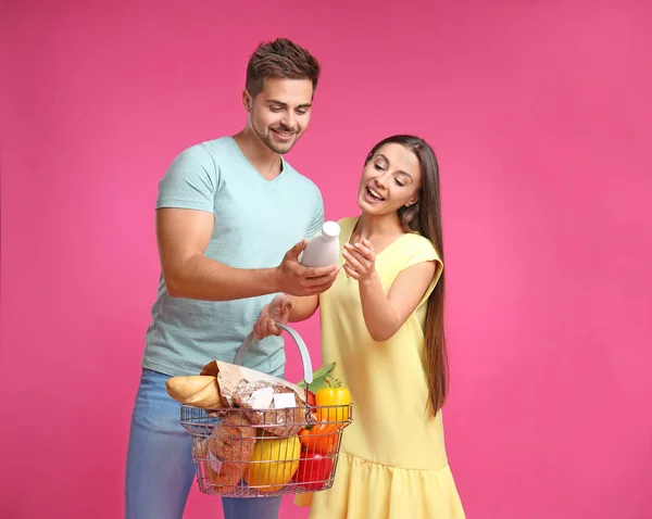 Casal jovem com cesta de compras cheia de produtos em fundo rosa — Fotografia de Stock