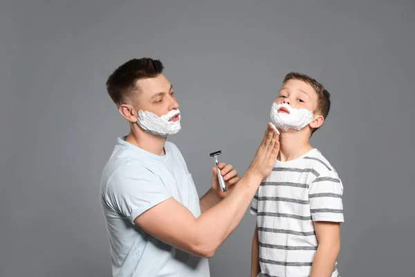 Tata stosujące piankę do golenia na twarzy syna, szare tło — Zdjęcie stockowe