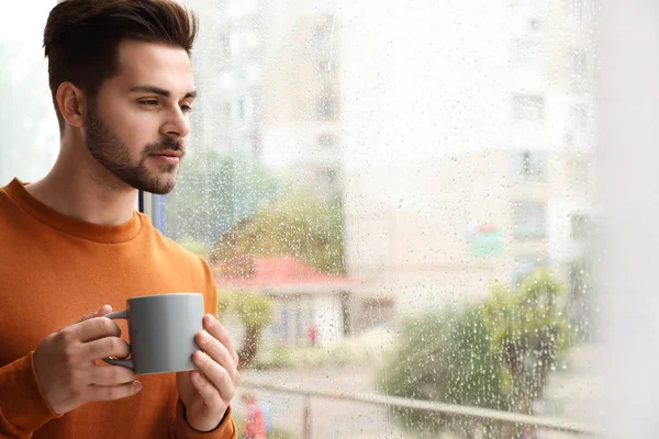 Thoughtful handsome man with cup of coffee near window indoors on rainy day — Stock Photo, Image