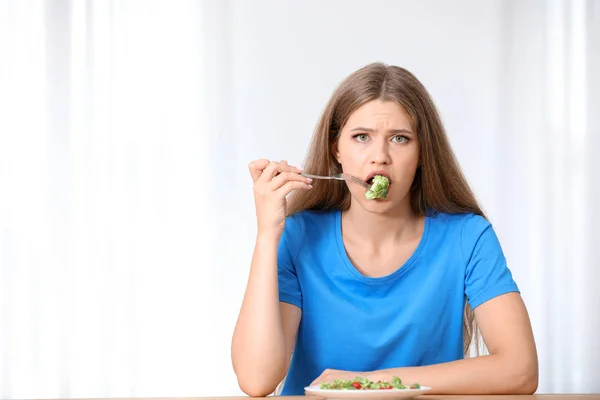 Portrait of unhappy woman eating broccoli salad at table on light background — Stock Photo, Image