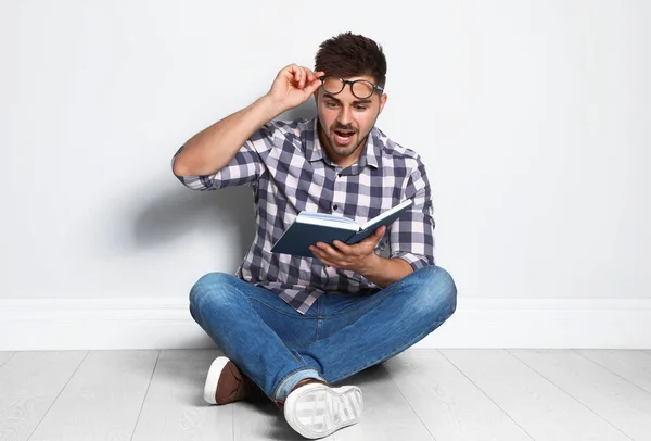 Knappe jonge man lezen boek op houten vloer in de buurt van lichte muur — Stockfoto