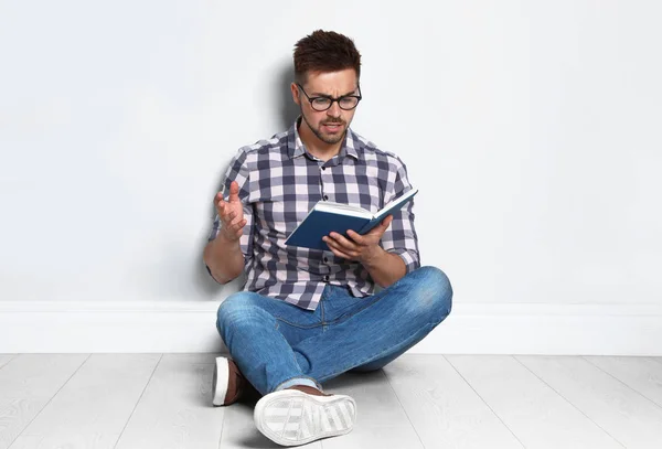 Knappe jonge man lezen boek op houten vloer in de buurt van lichte muur — Stockfoto