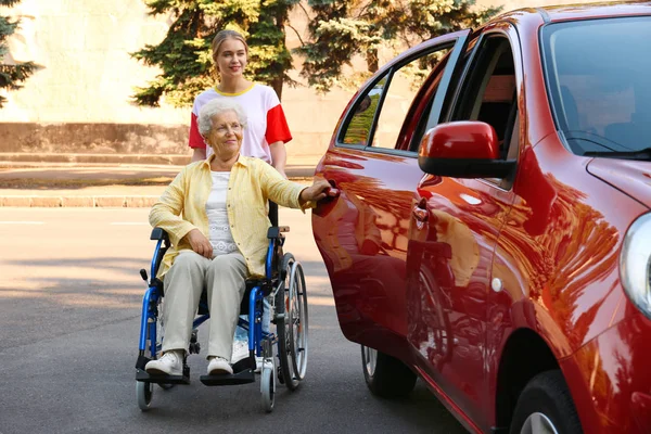 Jonge vrouw helpen gehandicapte grootmoeder in rolstoel te krijgen in de auto buitenshuis — Stockfoto