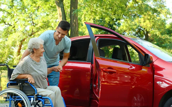 Молодой человек помогает пожилой женщине-инвалиду в инвалидном кресле садиться в машину на открытом воздухе — стоковое фото