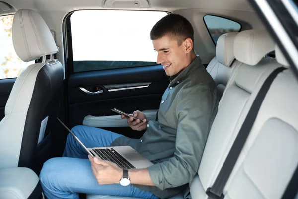 Atractivo joven que trabaja con el ordenador portátil y el teléfono inteligente en el asiento trasero en el coche de lujo — Foto de Stock