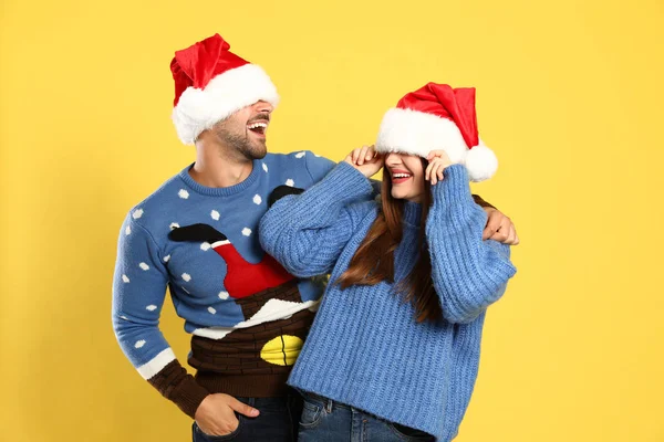 Coppia che indossa maglioni di Natale e cappelli di Babbo Natale su sfondo giallo — Foto Stock