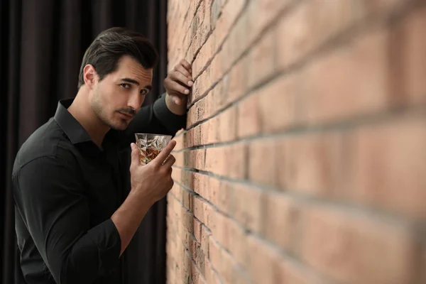 Hombre joven con vaso de whisky cerca de la pared de ladrillo en el interior. Espacio para texto —  Fotos de Stock