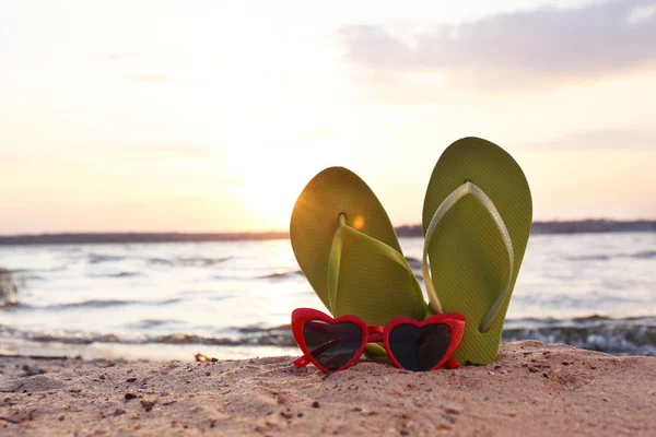 Stylish flip flops with sunglasses on sand near sea, space for text. Beach accessories — Stock Photo, Image