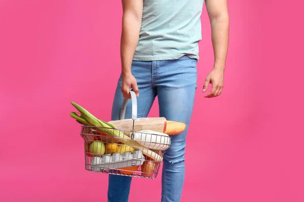 Mladý muž s nákupním košíkem plným produktů na růžovém pozadí, šatna — Stock fotografie