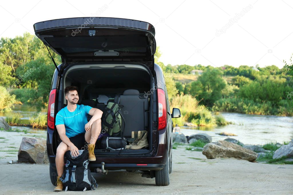 Young man near car with sleeping bag and camping equipment outdoors. Space for text
