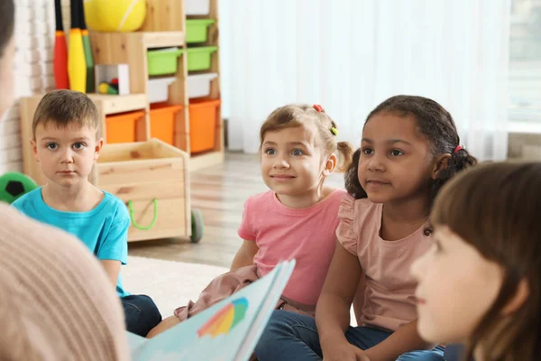Kindergarten teacher reading book to cute little children indoors — Stock Photo, Image