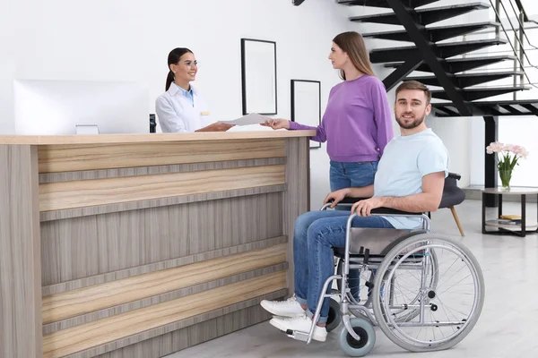 Frau mit Mann im Rollstuhl nahe Rezeptionistin in Klinik — Stockfoto