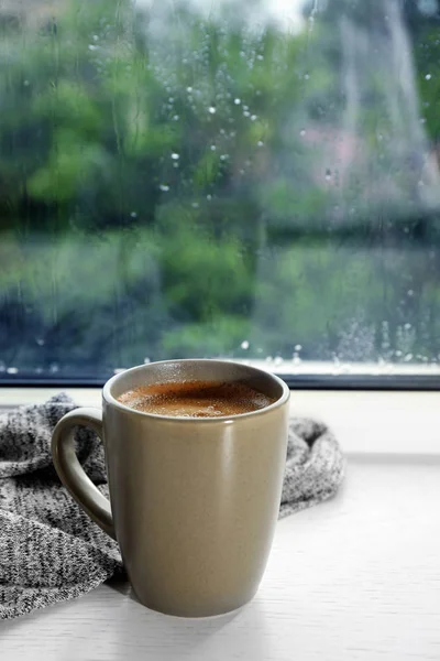 Copo de bebida quente no peitoril da janela contra vidro com gotas de chuva, espaço para texto — Fotografia de Stock