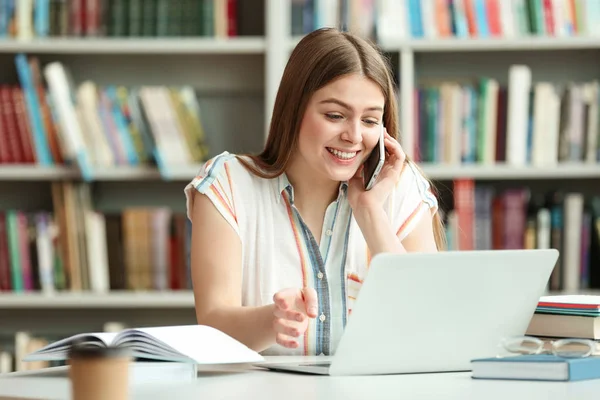 Jonge vrouw praten op telefoon en werken met laptop aan tafel in de bibliotheek — Stockfoto