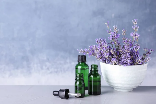 Bottiglie di olio essenziale e ciotola con fiori di lavanda su tavolo di pietra su sfondo blu. Spazio per testo — Foto Stock