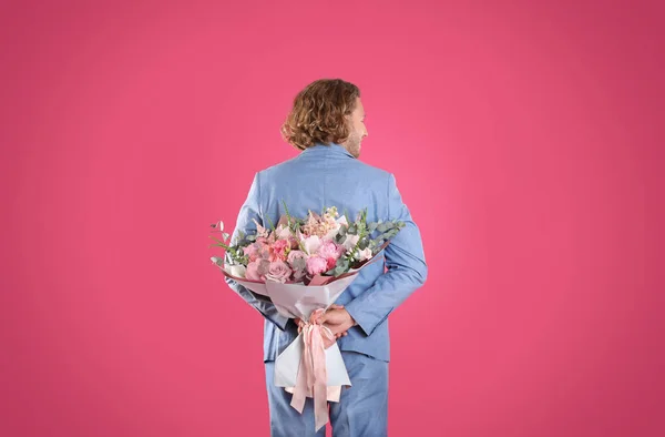 Uomo in abito elegante nascondendo bel mazzo di fiori dietro la schiena su sfondo rosa — Foto Stock