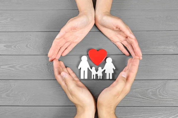 Young man and woman protecting family figure and red heart with their hands on grey wooden background, top view