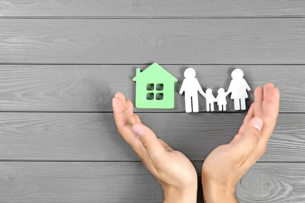 Young man protecting family figure and house model with his hands on grey wooden background, top view. Space for text