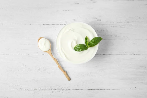 Bowl of fresh sour cream with basil and spoon on white wooden table, flat lay — Stock Photo, Image