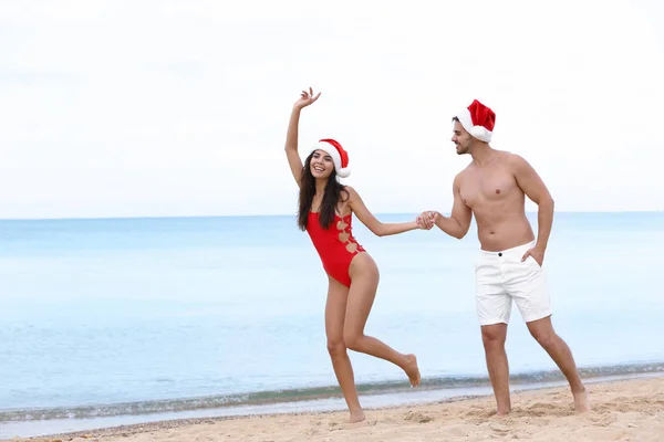 Feliz pareja joven con sombreros de Santa en la playa cerca del mar — Foto de Stock