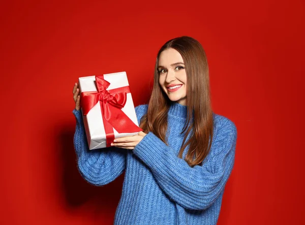 Νέα γυναίκα στο χριστουγεννιάτικο πουλόβερ κρατώντας κουτί δώρου σε κόκκινο φόντο — Φωτογραφία Αρχείου