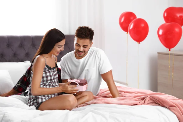 Jovem apresentando presente para sua namorada no quarto decorado com balões de ar. Celebração do Dia de São Valentim — Fotografia de Stock