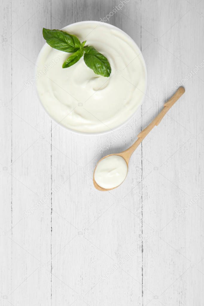 Bowl of fresh sour cream with basil and spoon on white wooden table, flat lay. Space for text