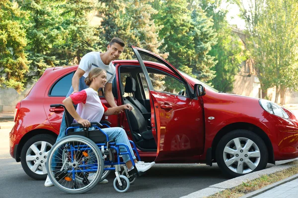 Молодой человек помогает женщине-инвалиду в инвалидном кресле садиться в машину на открытом воздухе — стоковое фото