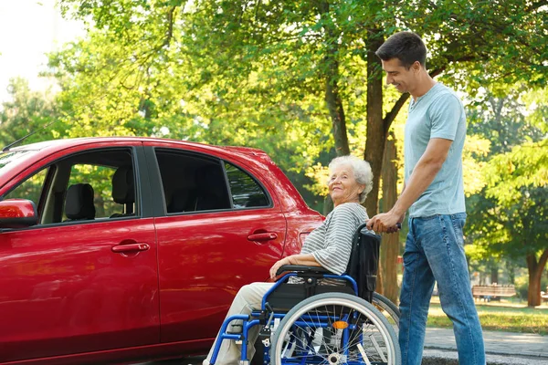 Młody mężczyzna z niepełnosprawnym starszym kobietą na wózku w pobliżu samochodu na zewnątrz — Zdjęcie stockowe