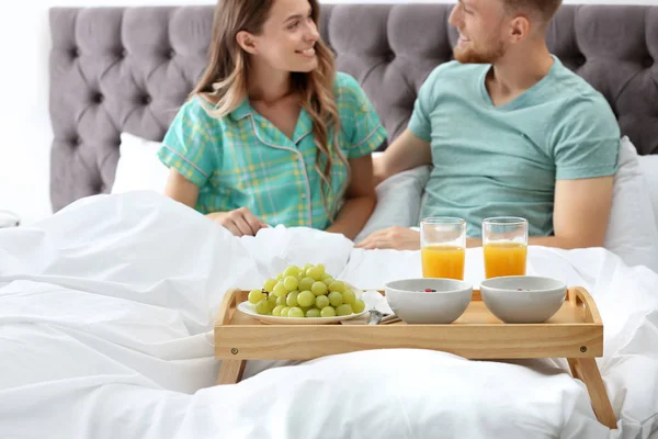 Bandeja con sabroso desayuno y pareja joven en la cama en casa — Foto de Stock