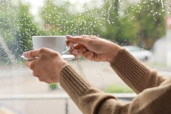 Młoda kobieta z filiżanką kawy w pobliżu okna wewnątrz w deszczowy dzień, zbliżenie — Zdjęcie stockowe