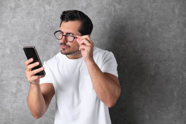 Gri arka planda cep telefonu kullanan gözlüklü genç bir adam. Görme sorunu