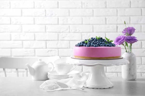 Stand with tasty blueberry cake and flowers on grey table against white brick wall. Space for text — Stock Photo, Image