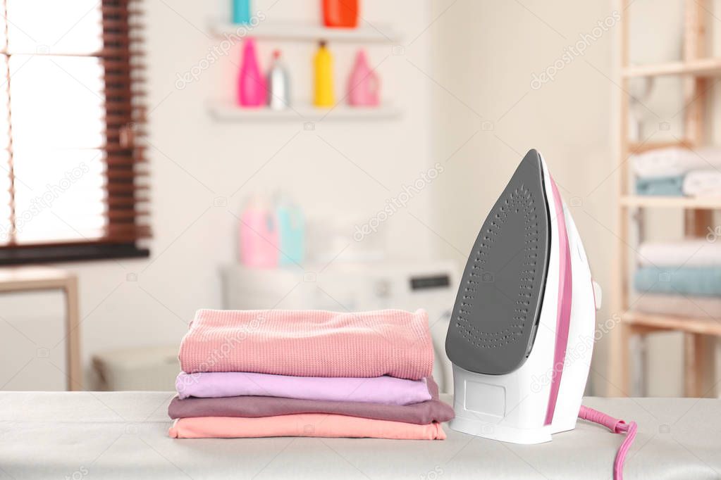 Modern electric iron and folded clothes on board in laundry room. Space for text