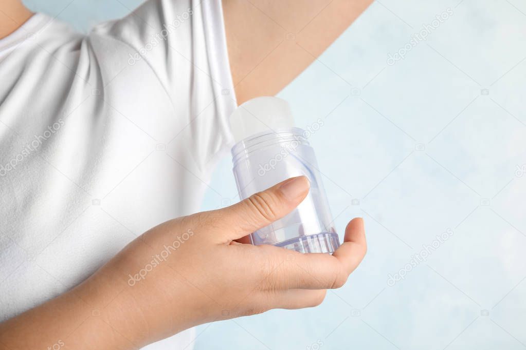 Young woman holding natural crystal alum deodorant near armpit on light blue background, closeup